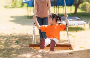 bambino con gioia oscillante su un' soleggiato giorno. contento giovane ragazza nel arancia camicia gode un' swing cavalcata nel il parco, con illuminata dal sole alberi nel il sfondo. foto