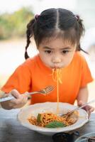 giovane ragazza bambini godendo un' forchettata di spaghetti. avvicinamento di un' poco ragazza nel un arancia camicia mangiare spaghetti con un' focalizzata espressione, all'aperto ristorante. foto