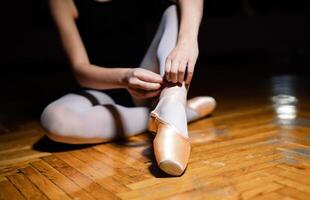 sconosciuto ballerina è legatura il nastro di pointe scarpe su il di legno pavimento nel un' balletto classe. il ballerina cravatte il pointes su snello gambe. avvicinamento foto