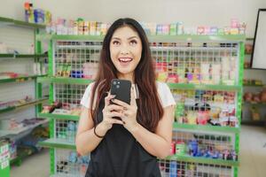 sorridente asiatico donna come un' cassiere è indossare nero grembiule e Tenere sua Telefono foto