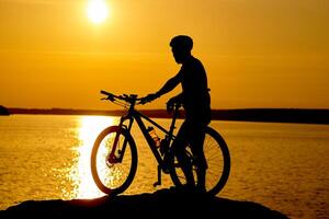 silhouette di sportivo equitazione un' bicicletta su il spiaggia. colorato tramonto nuvoloso cielo nel sfondo foto