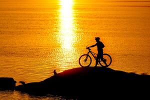 silhouette di sportivo equitazione un' bicicletta su il spiaggia. colorato tramonto nuvoloso cielo nel sfondo foto