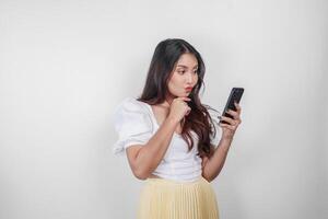 un' riflessivo asiatico donna è Tenere sua smartphone mentre immaginando sua pensieri, isolato di bianca sfondo. foto