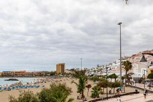 panoramico Visualizza di un' spiaggia ricorrere con ombrelloni, palma alberi, e turisti sotto un' nuvoloso cielo nel los cristiano, tenerife. foto