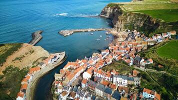 aereo Visualizza di un' costiero villaggio con dai tetti rossi case, un' porto con Barche, e scogliere prospiciente un' blu mare nel staithes, Inghilterra. foto