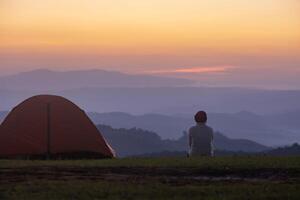 viaggiatore è seduta di il tenda durante durante la notte campeggio mentre guardare a il bellissimo panoramico tramonto al di sopra di il montagna per all'aperto avventura vacanza viaggio foto