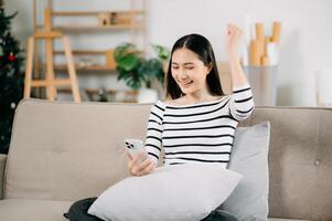 giovane asiatico donna rilassare su confortevole divano a casa sms messaggistica su smartphone e tavoletta sorridente ragazza uso cellulare shopping in linea a partire dal casa foto