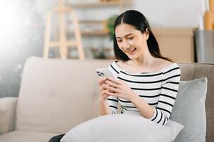 giovane asiatico donna rilassare su confortevole divano a casa sms messaggistica su smartphone e tavoletta sorridente ragazza uso cellulare shopping in linea a partire dal casa foto