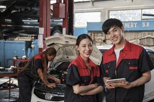 Due asiatico professionale meccanica, maschio ingegnere e compagno Sorridi e Guarda a telecamera, ispezionare riparazione opera lista di controllo a box auto, servizio auto Manutenzione, e fissaggio specialista occupazioni auto industria. foto