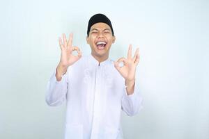 gioioso asiatico musulmano uomo mostrare ok cartello con ridendo espressione isolato su bianca sfondo foto