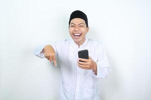estatico asiatico musulmano uomo ridendo mentre Tenere Telefono con puntamento dito giù isolato foto