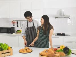 giovane bello uomo indossare occhiali e bellissimo donna Aiuto cucinando Pizza nel il bianca cucina felicemente. contento asiatico coppia mangiare Pizza nel il cucina. foto