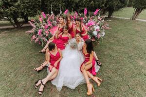 un' brunetta sposa e sua damigelle d'onore nel vestiti di il stesso colore sedersi con il sposa e rallegrarsi con fiori nel loro mani vicino il solenne arco. nozze nel natura. foto