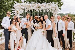 il sposa e sposo e loro amici posa vicino il arco. lungo treno di il vestire. elegante nozze. estate nozze nel natura foto