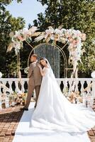 un' sposo nel un' completo da uomo e un' sposa nel un' nozze vestito In piedi vicino un arco fatto di fiori durante un' nozze cerimonia. estate nozze nel natura. foto