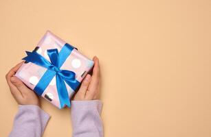 un' donna di mano detiene un' regalo scatola avvolto nel un' blu seta nastro su un' beige sfondo, superiore Visualizza foto