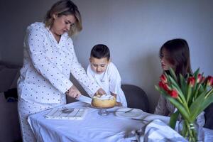 famiglia di tre, madre, adolescenziale figlia e poco figlio, mangiare torta nel pigiama a un' tavolo con tulipani foto