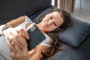 giovane donna bugie su il divano e legge un' in linea libro su sua smartphone foto