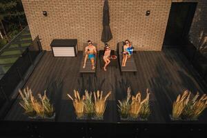 un' contento famiglia nel costumi da bagno prendere il sole nel estate su loro terrazza su sole letti foto