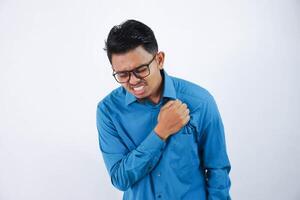 asiatico giovane uomo con bicchieri Tenere il suo il petto nel il petto dolore o coronario cuore malattia indossare blu camicia isolato su bianca sfondo foto