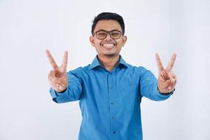 positivo o contento asiatico uomo con bicchieri a forma di v dito gesto o pace indossare blu camicia isolato su bianca sfondo foto