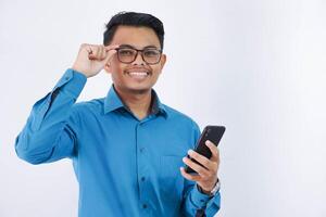 sorridente asiatico dipendente uomo Tenere Telefono e Tenere il bordo di il bicchieri indossare blu camicia isolato su bianca sfondo foto