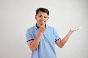 sorridente asiatico uomo con un Aperto mano il lato e mentre Tenere il mento indossare blu t camicia isolato su bianca sfondo foto