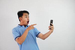 ritratto asiatico uomo scioccato Tenere Telefono e puntamento a il Telefono con un' dito indossare blu polo t camicia isolato su bianca sfondo foto