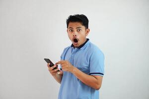 ritratto asiatico uomo scioccato Tenere Telefono e puntamento a il Telefono con un' dito indossare blu polo t camicia isolato su bianca sfondo foto