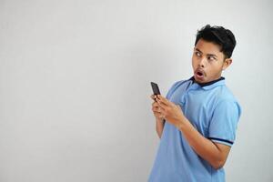 scioccato giovane asiatico uomo Tenere Telefono e Aperto bocca indossare blu t camicia isolato su bianca sfondo foto