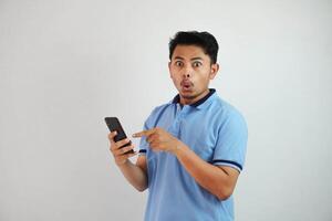 ritratto asiatico uomo scioccato Tenere Telefono e puntamento a il Telefono con un' dito indossare blu polo t camicia isolato su bianca sfondo foto