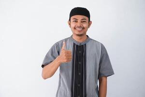 sorridente o contento giovane musulmano uomo mostrando mano pollici su indossare grigio musulmano Abiti isolato su bianca sfondo foto