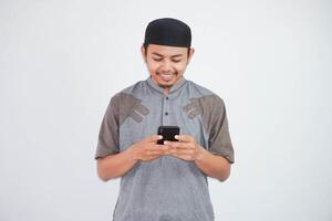 contento o sorridente giovane asiatico musulmano uomo Tenere Telefono cellulare indossare koko Abiti isolato su bianca sfondo. Ramadan concetto foto