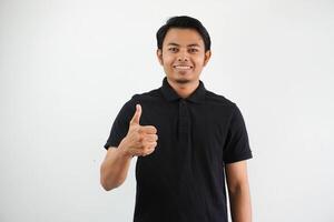 ritratto di sorridente giovane asiatico uomo dare pollice su indossare nero polo t camicia isolato su bianca sfondo foto