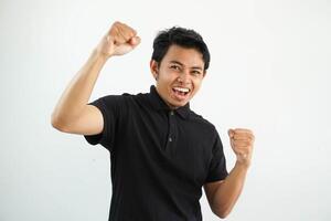 sorridente o contento giovane asiatico uomo stretto il suo pugni applauso spensierato e emozionato. vittoria concetto, indossare nero polo t camicia isolato su bianca sfondo foto