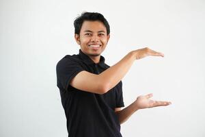 giovane asiatico uomo in posa su un' bianca fondale sorridente e felicità Tenere un' copia spazio fra mani, indossare nero polo t camicia. foto