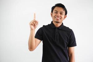sorridente giovane asiatico uomo puntamento su con contento viso espressione indossare nero polo t camicia isolato su bianca sfondo foto