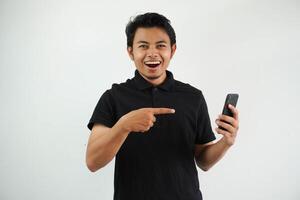 eccitato giovane asiatico uomo sorridente contento mentre puntamento per mobile Telefono quello lui hold indossare nero polo t camicia isolato su bianca sfondo foto