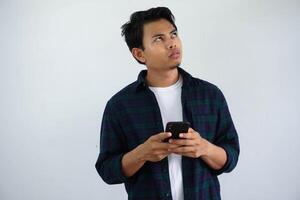 giovane asiatico uomo pensare qualcosa mentre digitando su il suo mobile Telefono isolato su bianca sfondo. foto
