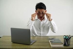 sorpreso giovane asiatico uomo d'affari nel un' posto di lavoro Aperto bocca con Tenere bicchieri indossare bianca camicia isolato foto