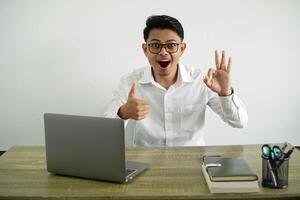 giovane asiatico uomo d'affari nel un' posto di lavoro mostrando ok cartello e pollice su gesto, indossare bianca camicia con bicchieri isolato foto