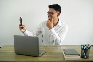 giovane asiatico uomo d'affari nel un' posto di lavoro fabbricazione un' autoscatto con mobile Telefono, indossare bianca camicia con bicchieri isolato foto
