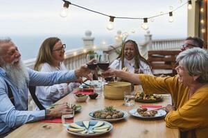 multirazziale anziano amici avendo divertimento cenare insieme e tostatura con rosso vino su Casa terrazza cena - cibo e vacanze concetto foto