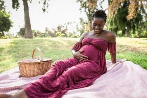 africano donna carezzevole sua incinta pancia mentre fare un' picnic nel parco - maternità stile di vita concetto foto