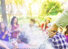 gruppo di persone avendo un' barbecue e potabile rosso vino nel un' parco - vicino su di maschio mano grigliate carne - contento amici avendo divertimento con un' picnic - concetto di le persone, amicizia e divertimento foto