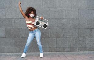 giovane afro donna indossare viso maschera danza all'aperto mentre ascoltando per musica con senza fili cuffie e Vintage ▾ Boombox stereo foto