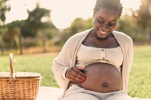 africano donna carezzevole sua incinta pancia mentre fare un' picnic nel parco - maternità stile di vita concetto foto