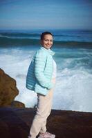 contento sorridente donna in piedi di oceano su il roccioso scogliera, guardare a telecamera, godendo il Visualizza di bellissimo onde foto