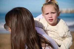 bellissimo bambino ragazza guardare a telecamera mentre sua amorevole madre trasporto sua su sua Indietro, a piedi insieme su il spiaggia foto