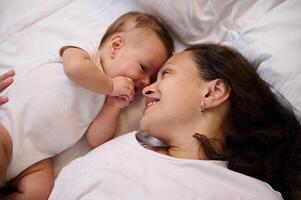 avvicinamento ritratto di un' contento sorridente madre con sua carino poco bambino dire bugie su il letto, sensazione emotivo contatto foto